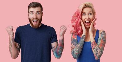 Indoor photo of joyful young pretty blue-eyed couple with tattooes going out of mind from happiness and looking exitedly to camera, standing over pink background in casual shirts