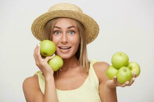 Portrait of young beautiful cheerful blue-eyed blonde female with natural makeup in straw boater hat looking excitedly at camera with joyful smile, isolated over white background with green apples photo