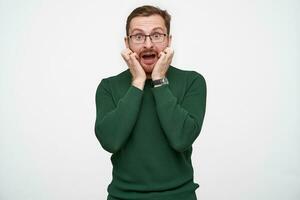 Studio shot of frightened young brunette male with beard wearing eyewear and green sweater while standing over white background, rounding scaredly eyes with opened mouth photo