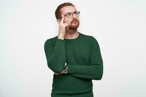 Studio shot of pensive young brunette bearded male in green pullover holding forefinger on his temple and looking thoughtfully aside with folded lips, isolated over white background photo