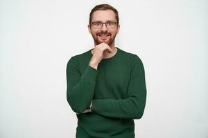 Indoor photo of happy young brunette bearded guy in eyewear leaning his chin on raised hand and smiling cheerfully to camera, wearing green pullover against white background