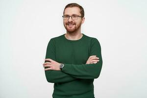 Indoor photo of positive good looking young bearded man in glasses wearing green sweater while standing over white background, smiling slightly and keeping hands folded