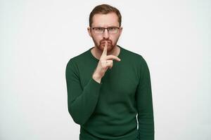 Indoor photo of young brunette bearded guy in glasses raising hand with hush gesture while posing over white background, frowning eyebrows and looking seriously at camera