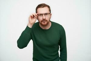 Doubting young brunette short haired pretty male with beard looking attentively at camera and holding hand on his eyewear, posing over white background in casual clothes photo