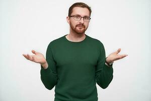 Puzzled young handsome brown haired bearded male shugging with raised palms and looking at camera with folded lips, wearing green pullover and eyewear over white background photo