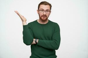 Confused young brunette man in glasses with short haircut raising emotionally hand and wrinkling forehead while looking at camera, isolated over white background photo