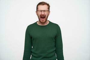 Stressed young short haired brunette man with beard wearing eyewear and green sweater posing over white background with hands down, screaming desperately with closed eyes photo
