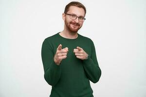 Positive young lovely brunette bearded man with short haircut pointing to camera with raised forefingers and smiling nicely, wearing green sweater and glasses over white background photo