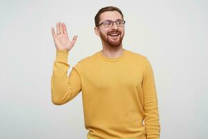 Shot of cheerful young handsome brown haired bearded guy in eyewear raising palm in hello gesture while looking positively aside with sincere smile, posing over white background in mustard pullover photo