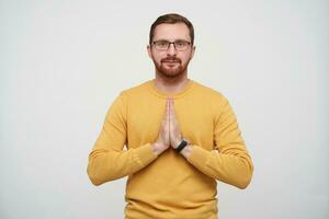 retrato de hermoso joven barbado masculino con corto Corte de pelo mirando a cámara con calma cara y plegable palmas en namaste gesto, posando terminado blanco antecedentes en mostaza pull-over foto