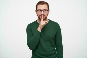 Studio shot of handsome young bearded male in glasses with brown short hair holding forefinger on his lips and looking aside, asking to keep secret, isolated over white background photo