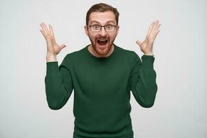 Overjoyed young pretty brunette bearded male in glasses raising happily hands and looking at camera with wide eyes and mouth opened, standing against white background photo