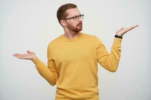 Puzzled young brunette bearded man in glasses frowning eyebrows and looking at his raised palm while posing over white background, wearing casual clothes and wristwatch photo