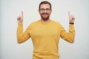 Portrait of happy young brunette bearded guy in eyewear pointing upwards with raised forefingers and smiling widely at camera, dressed in mustard pullover over white background photo