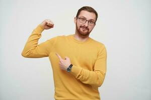 Positive young attractive brown haired male with beard wearing eyewear and mustard pullover while standing over white background, showing on his biceps with index finger and smiling slightly at camera photo
