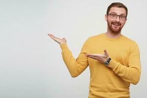 Studio shot of cheerful young handsome brunette male with beard showing aside with raised palms and looking happily at camera, dressed in mustard sweater and eyewear over white background photo