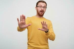 Scared young bearded male with brown short hair keeping raised hands in front of him and rounding frightened eyes while looking to camera, isolated over white background photo