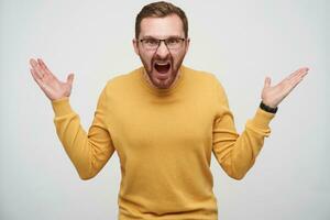 Studio photo of angry young brown haired bearded man in glasses raising emotionally palms up and shouting heatedly at camera, standing against white background