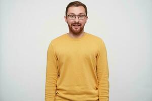 Suprised young brown haired pretty guy with beard wearing casual pullover and glasses while standing over white background with hands down, looking surprisedly at camera photo