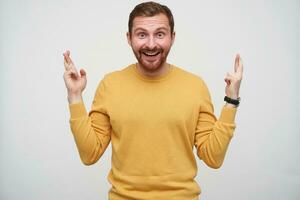 Overjoyed attractive young bearded man with brown short hair looking at camera with wide eyes and mouth opened and crossing his fingers for good luck, standing against white background photo