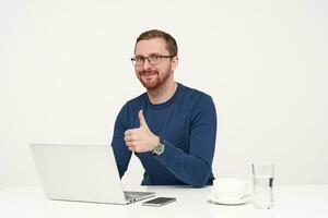 Studio shot of young positive fair-haired male in glasses showing raised thumb and smiling slightly at camera while sitting at table over white background photo