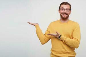 Indoor photo of joyful pretty young brunette bearded man with short haircut pointing aside with palms and raising happily eyebrows while looking at camera, isolated over white background