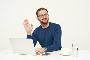 Handsome young unshaved fair-haired man dressed in blue sweater raising palm in hello gesture while looking aside, working with his laptop over white background photo