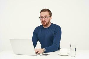 Confused young fair-haired man in eyewear keeping hands on keyboard and looking surprisedly on screen of his laptop while sitting over white background photo