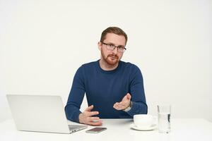 Bewildered young pretty bearded guy in eyewear looking confusedly at camera and raising perplexedly palm, isolated over white background in blue pullover photo