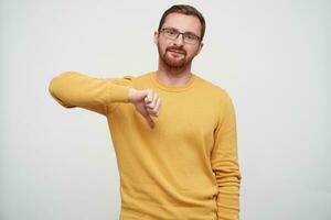 Studio photo of handsome young brunette bearded guy in glasses showing down with thumb and looking at camera with folded lips, isolated over white background