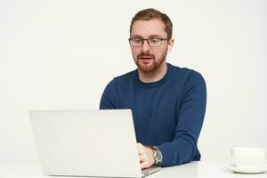 Indoor shot of young fair-haired man in glasses looking seriously at his laptop while typing text on keyboard, being concentrated on his work while posing isolated over white background photo