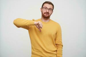 Shot of displeased young bearded male with brown short hair twisting his mouth with pout and pointing with thumb down, wearing mustard sweater while standing over white background photo