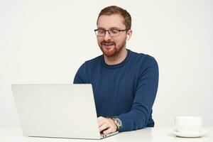 Positive young pretty bearded male with earphones keeping hands on keyboard and smiling gladly while typing text, isolated against white background photo