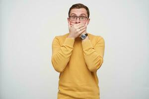 Shocked young pretty bearded male in glasses with brown short hair covering his mouth with raised palms and looking amazedly at camera, isolated over white background photo