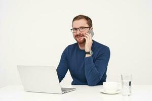 Indoor shot of young bearded male in glasses keeping mobile phone in raised hand while having pleasant talk and looking surprisedly at camera, isolated over white background photo