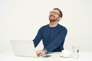 Joyful young bearded fair-haired man throwing back his head while laughing and keeping eyes closed, hearing funny joke while working with his laptop over white background photo