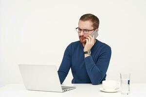 Concerned young unshaved fair-haired businessman in glasses making call with his smartphone while working over white background in casual clothes photo