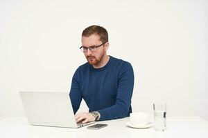 Indoor shot of young concentrated bearded guy in glasses looking seriously on screen of his laptop while working and keeping hands on keyboard, isolated over white background photo