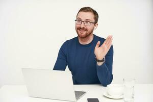 Cheerful young handsome unshaved fair-haired man smiling gladly while raising hand in hello gesture, sitting at table over white background with laptop photo
