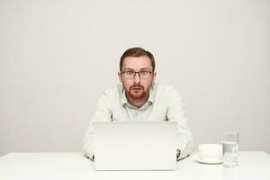 Agitated young short-haired bearded male in glasses keeping his hands on keyboard of laptop while looking surprisedly at camera, isolated over white background photo