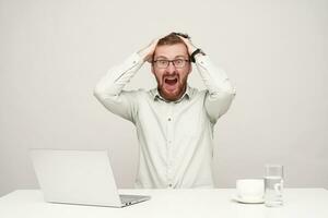 Bemused young handsome bearded male in eyewear clutching his head wit raised hands while looking amazedly at camera with stressed face, isolated over white background photo