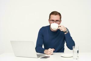 Studio shot of young bearded male in glasses having coffee break while working with his laptop and looking at camera, dressed in blue sweater while sitting over white background photo