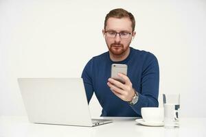 Studio photo of young fair-haired male in glasses keeping smartphone in raised hand and looking seriously on screen while reading message, posing over white background