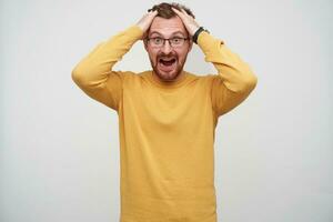 Excited young brunette attractive male with beard and short haircut clutching his head and looking at camera with wide eyes and mouth opened, dressed mustard pullover over white background photo