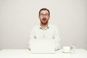 Bemused young pretty bearded man dressed in white shirt looking surprisedly at camera with wide mouth opened while typing text on his laptop, isolated over white background photo