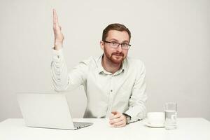 Studio shot of young bearded guy dressed in formal clothes keeping his lips folded while looking at camera and raising up hand, sitting over white background photo