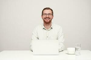 Happy young handsome unshaved male in eyewear looking gladly at camera with wide smile while working with his laptop over white background, being in high spirit photo