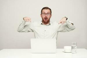 Dazed young handsome unshaved fair-haired man showing amazedly on his laptop with forefingers while looking at camera with wide eyes opened, posing over white background photo