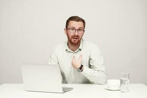 Puzzled young handsome unshaved fair-haired man looking confusedly at camera and grimacing his face while sitting at table with laptop over white background photo