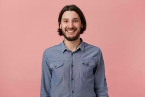 Photo of smiling bearded young man with long combed dark hair in denim shirt, looking at the camera isolated over pink background.